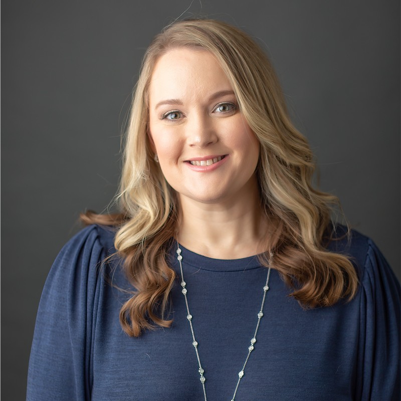 Smiling blonde woman in a navy blouse, posed against a gray studio backdrop.