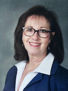 Portrait of a smiling woman with dark hair and glasses, wearing a blue blazer and white shirt against a neutral backdrop.